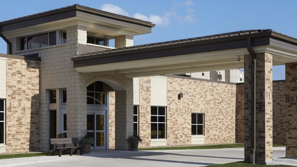 A modern brick building with large windows and a covered entrance. There are two potted plants and a bench near the glass doors, and the sky is clear with a few clouds.