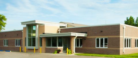 A single-story brick building with large windows and a small entrance canopy, surrounded by a paved area and some greenery, under a partly cloudy sky.