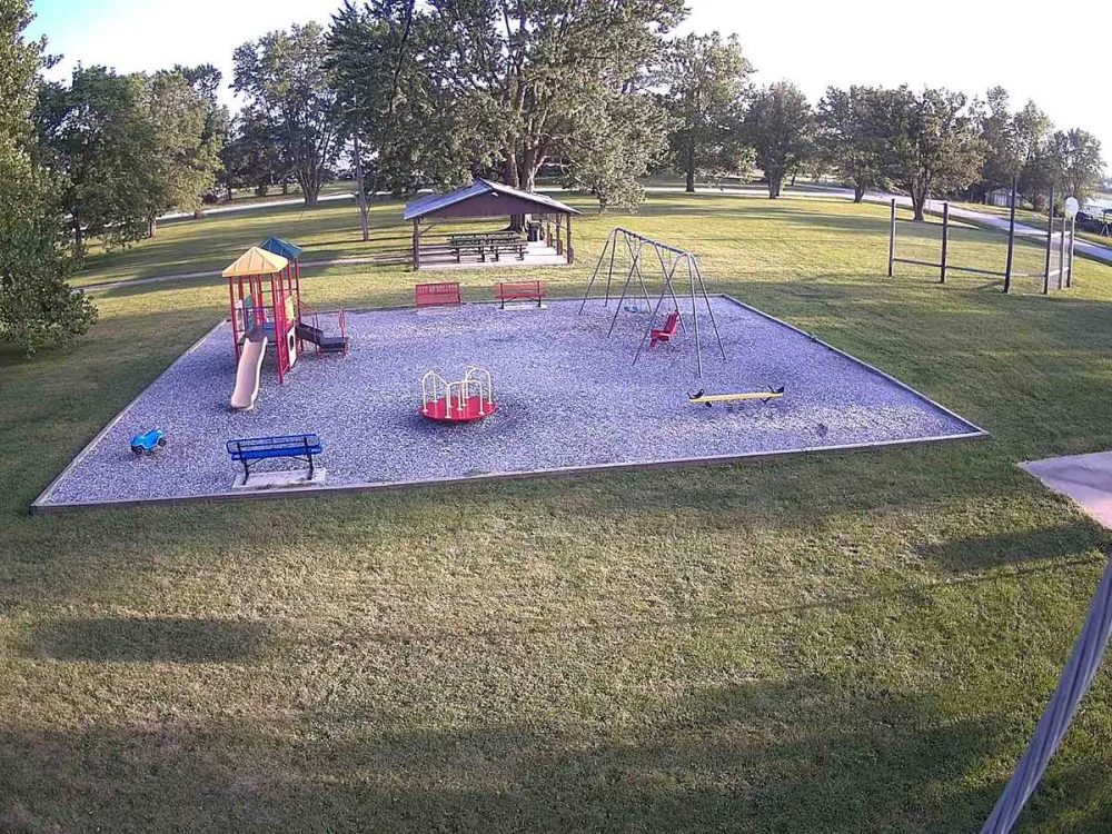 A playground with a slide, swings, a merry-go-round, and a seesaw sits on a gravel area surrounded by grass, trees, benches, and a picnic shelter in a park.