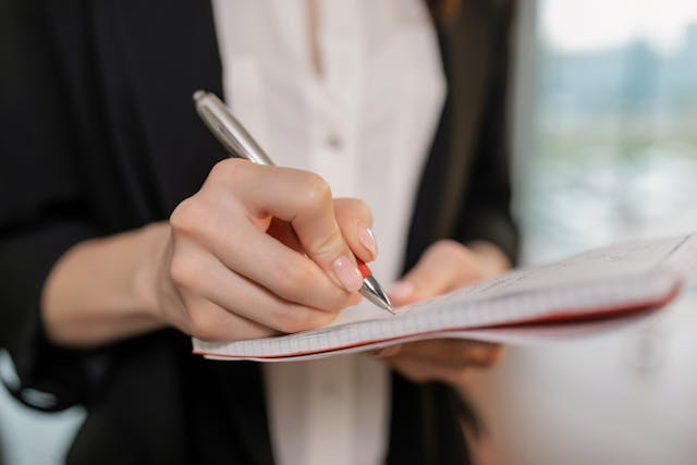 Close-up of a person writing in a notebook with a pen. The individual is wearing a white shirt and a dark jacket, and the background is softly blurred.