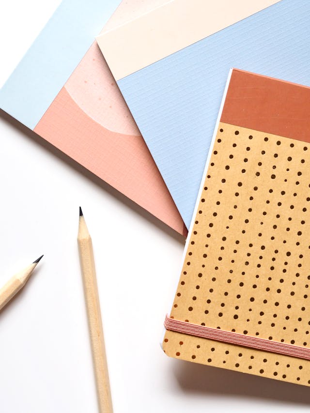 Three notebooks with pastel covers and geometric patterns are stacked on a white surface, next to two sharpened wooden pencils. One notebook has a brown cover with small black dots and a pink elastic band.