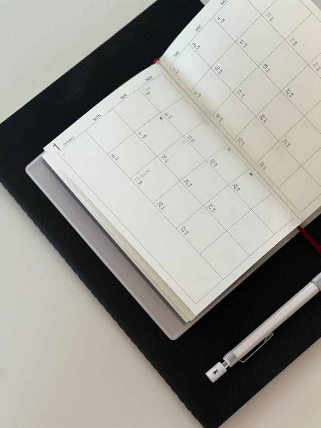 A monthly planner lies open on a desk with a pen beside it and a closed laptop underneath. The calendar pages are blank, ready to be filled in.
