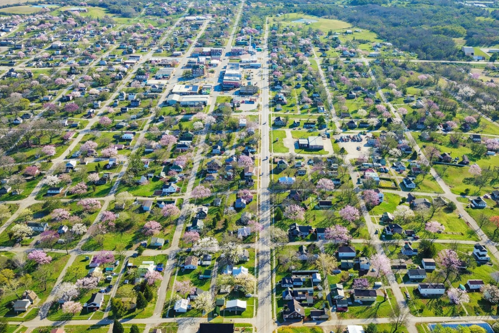 Aerial view of a suburban neighborhood with tree-lined streets, green lawns, and blooming trees. Houses are evenly spaced, and roads form a grid pattern. Commercial buildings are visible in the upper center of the image.