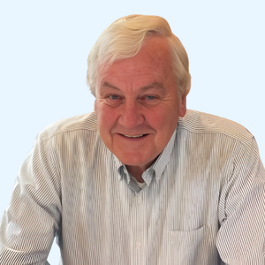 An older man with gray hair smiles while wearing a light-colored, striped button-up shirt against a plain, light background.