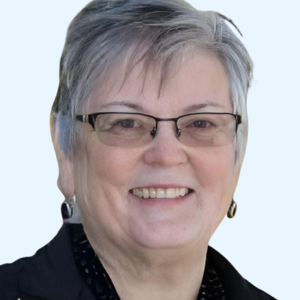 Smiling older woman with short gray hair, wearing glasses, earrings, and a black jacket, posing in front of a light background.