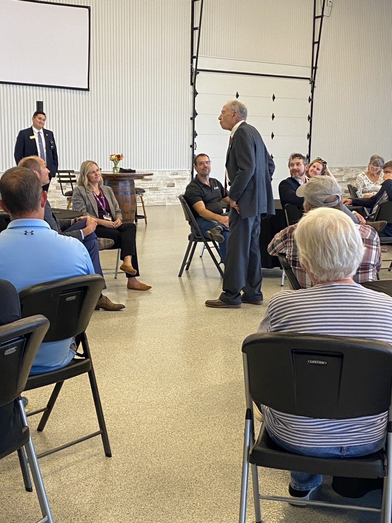 An older man in a suit stands and speaks to a seated audience in a large, bright room. People are sitting in chairs arranged in a circle, listening attentively. A woman and two men sit at the front near a barrel-shaped table.